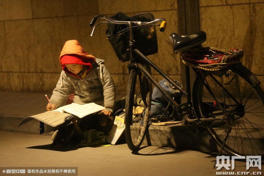 鄭州中原路京廣鐵路大橋下涵洞的馬路牙子邊，一位戴著紅色辣椒帽的小男孩趴在一個小馬扎上，上面鋪著一張車墊，在微弱的燈光下寫作業(yè)