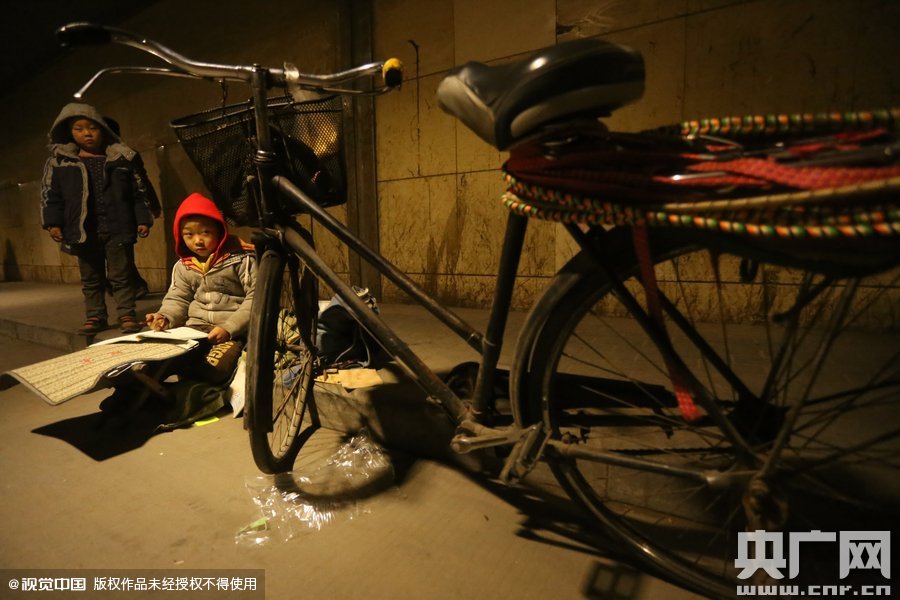 2015年11月8日晚，鄭州，7歲男孩涵洞橋下陪父母練攤，路邊燈下坐小馬扎上寫作業(yè)。